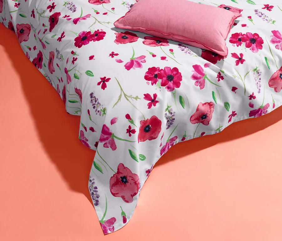 Eine weiße Renforcé-Multifunktions-Überwurf mit Blumenmuster und ein rosa Kissen auf korallefarbenem Hintergrund.