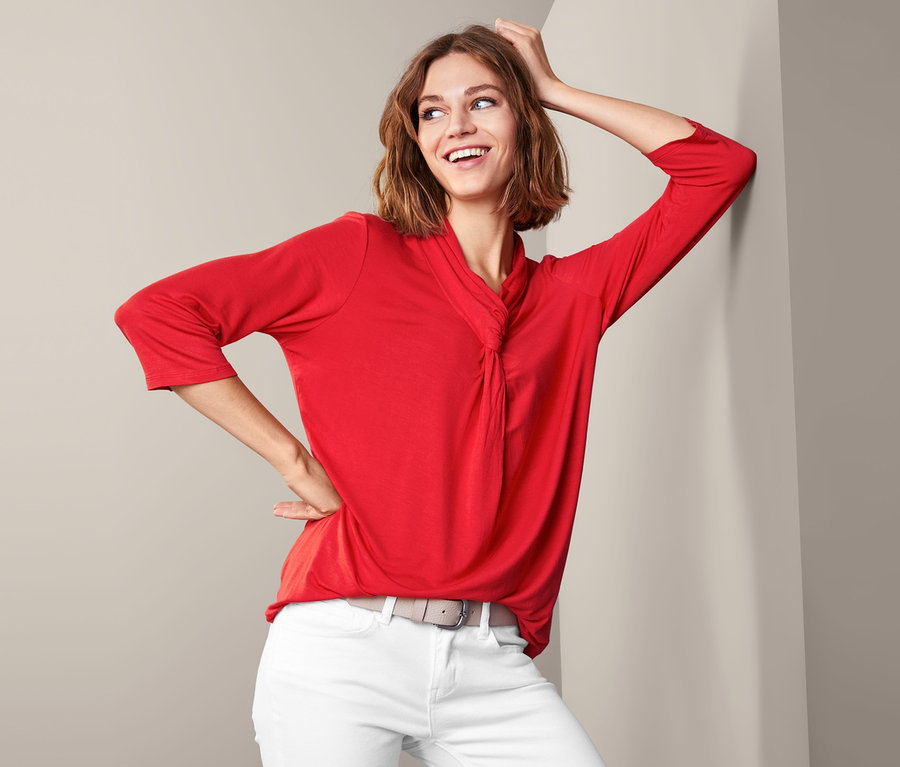 Frau lehnt an der Wand und trägt ein rotes Shirt mit Knotendetail, weiße Hose und grauen Gürtel.