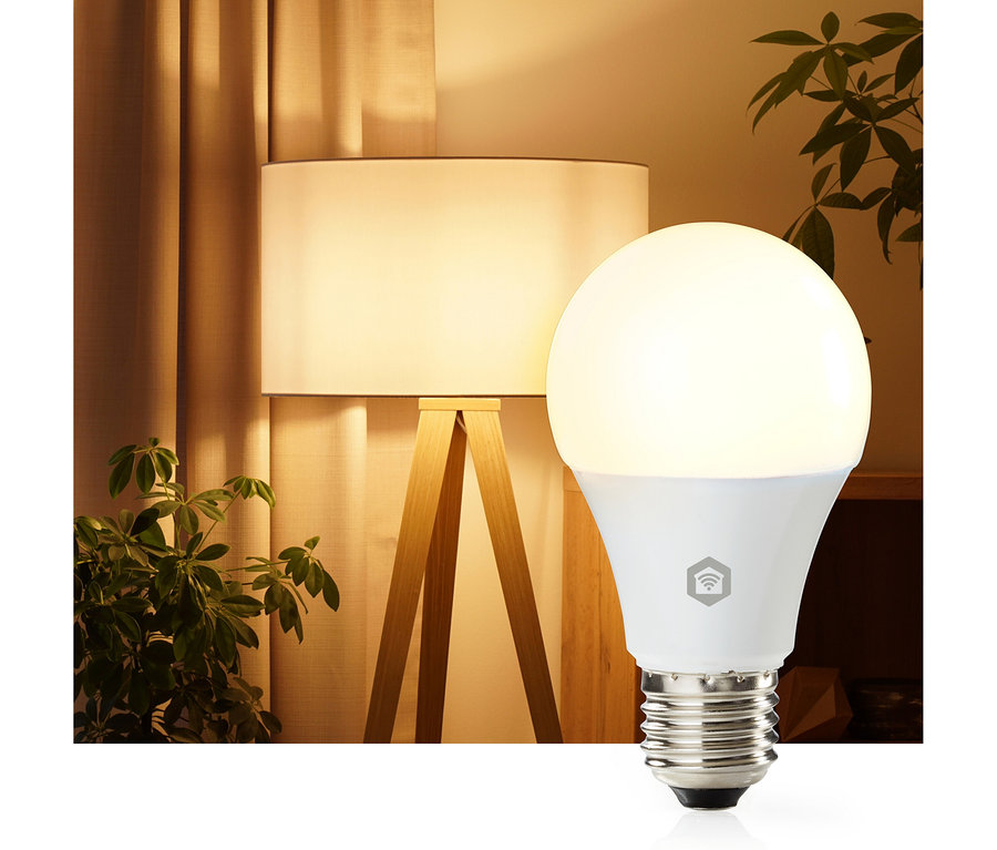 Eine leuchtende weiße Smart-Glühbirne mit sichtbarem Schraubsockel und Smart-Home-Symbol, vor einem warm beleuchteten Raum mit Stehlampe, hellen Vorhängen und einer grünen Pflanze.