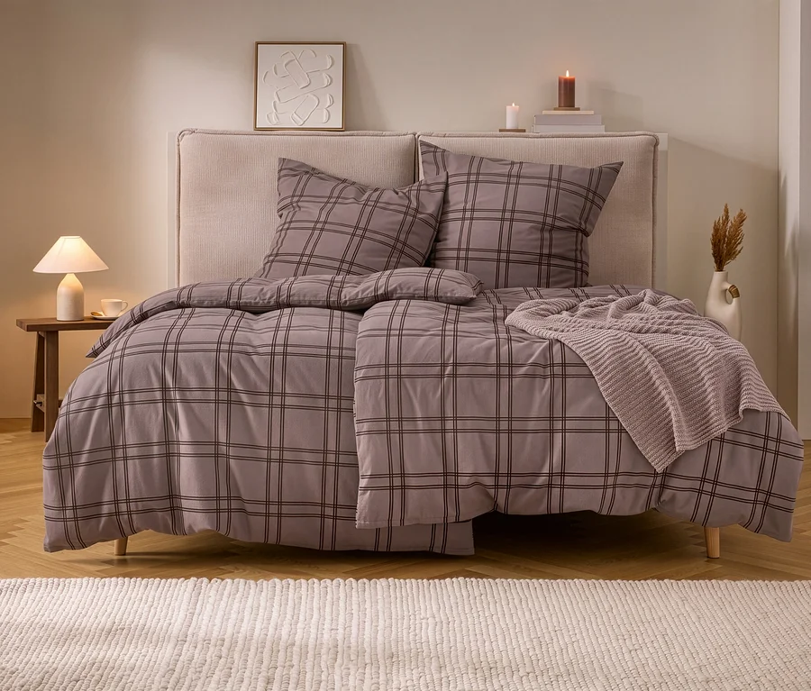 Doppelbett mit karierter Biber-Bettwäsche in Grau-Braun und beigem Stricküberwurf. Daneben ein Nachttisch mit Lampe und Kerzen.