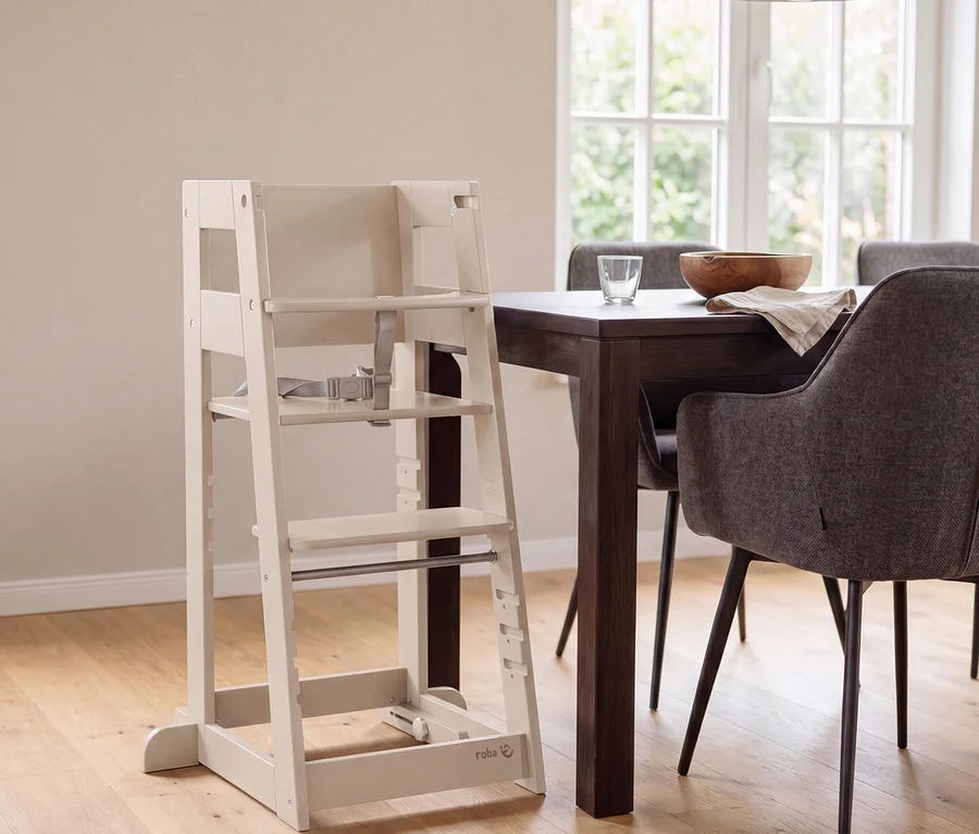 Ein heller Hochstuhl aus Holz mit grauem Gurt steht neben einem dunklen Esstisch, auf dem eine Holzschale, ein Glas und eine Serviette liegen. Graue Polsterstühle umgeben den Tisch, im Hintergrund ist ein Fenster zu sehen.