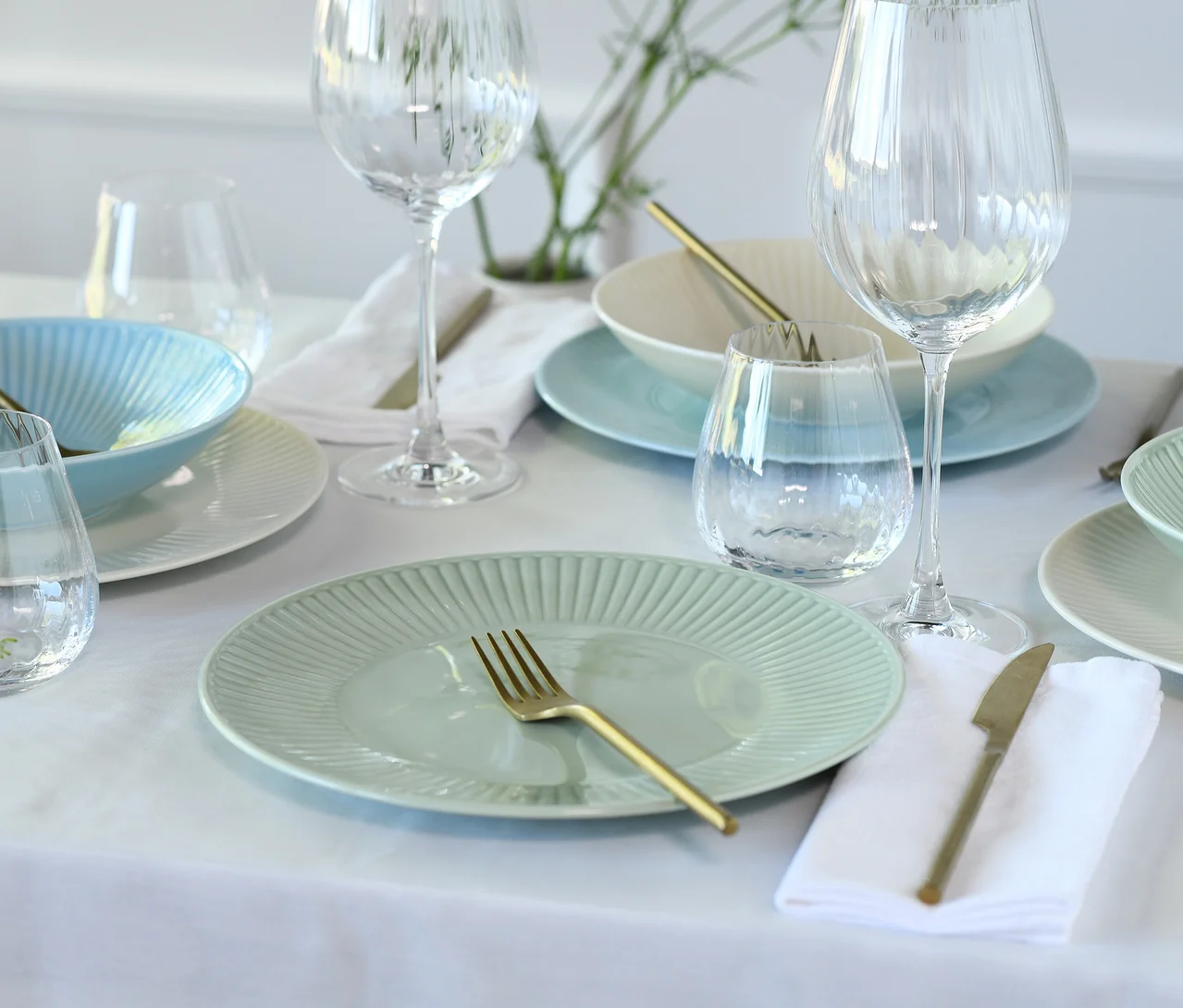 Ein gedeckter Tisch mit einem creaTable Kombiservice »Valencia Pastell« in Mint, Hellblau und Weiß, goldenem Besteck und Gläsern auf einer weißen Tischdecke.
