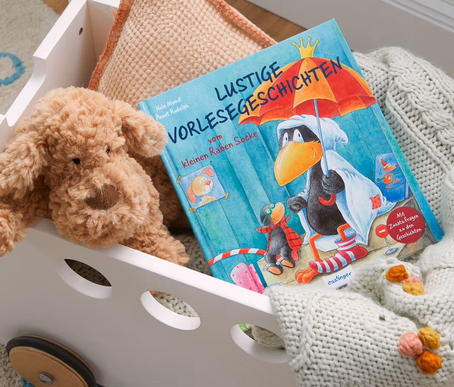In einem weißen Spielzeugwagen liegen ein Plüschhund neben dem Buch "Lustige Vorlesegeschichten vom kleinen Raben Socke", einem beigen Kissen und einer Strickdecke.