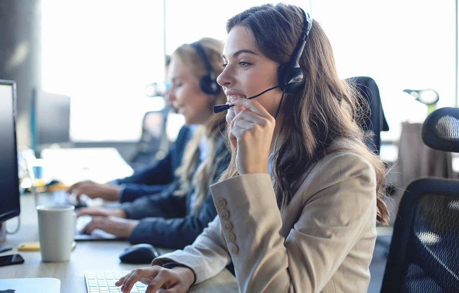 Eine Frau mit Headset spricht und tippt in einem Büro auf einer Tastatur. Im Hintergrund sind weitere Personen.