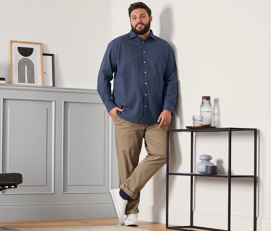 Ein Mann in einem Hemd mit Comfort Fit, einer camelfarbenen Chino und dunkelblauen Merinosocken (4 Paar, dunkelblau, rot und dunkelgrün) steht in einem Raum.