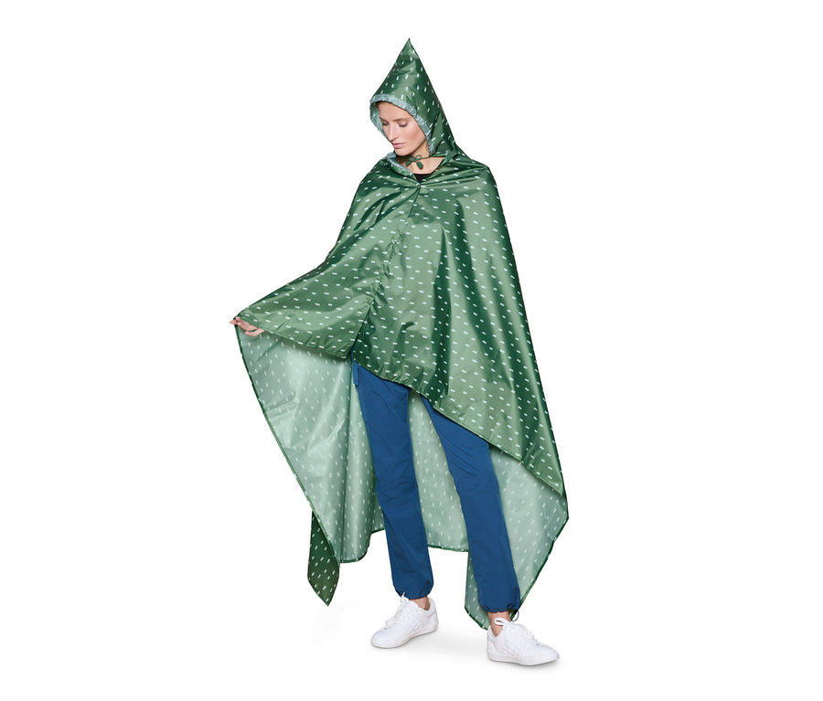 Frau posiert in einer grünen 2-in-1-Picknickdecke mit Kapuze und weißem Muster, trägt blaue Hosen und weiße Turnschuhe.