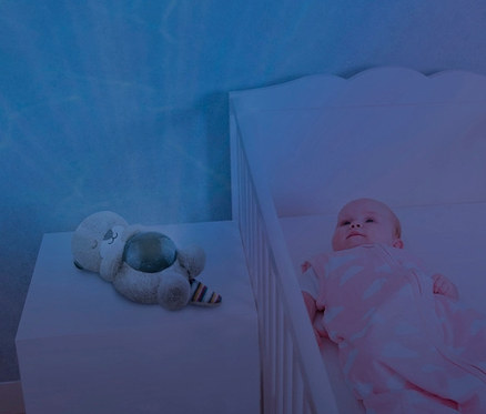 Baby liegt im Gitterbett neben ZAZU-Ozeanprojektor »Otto«.
