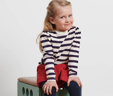 Mädchen sitzt auf grünem Spielzeug, trägt Kinder-Langarmshirt, roten Rock und Stiefel.