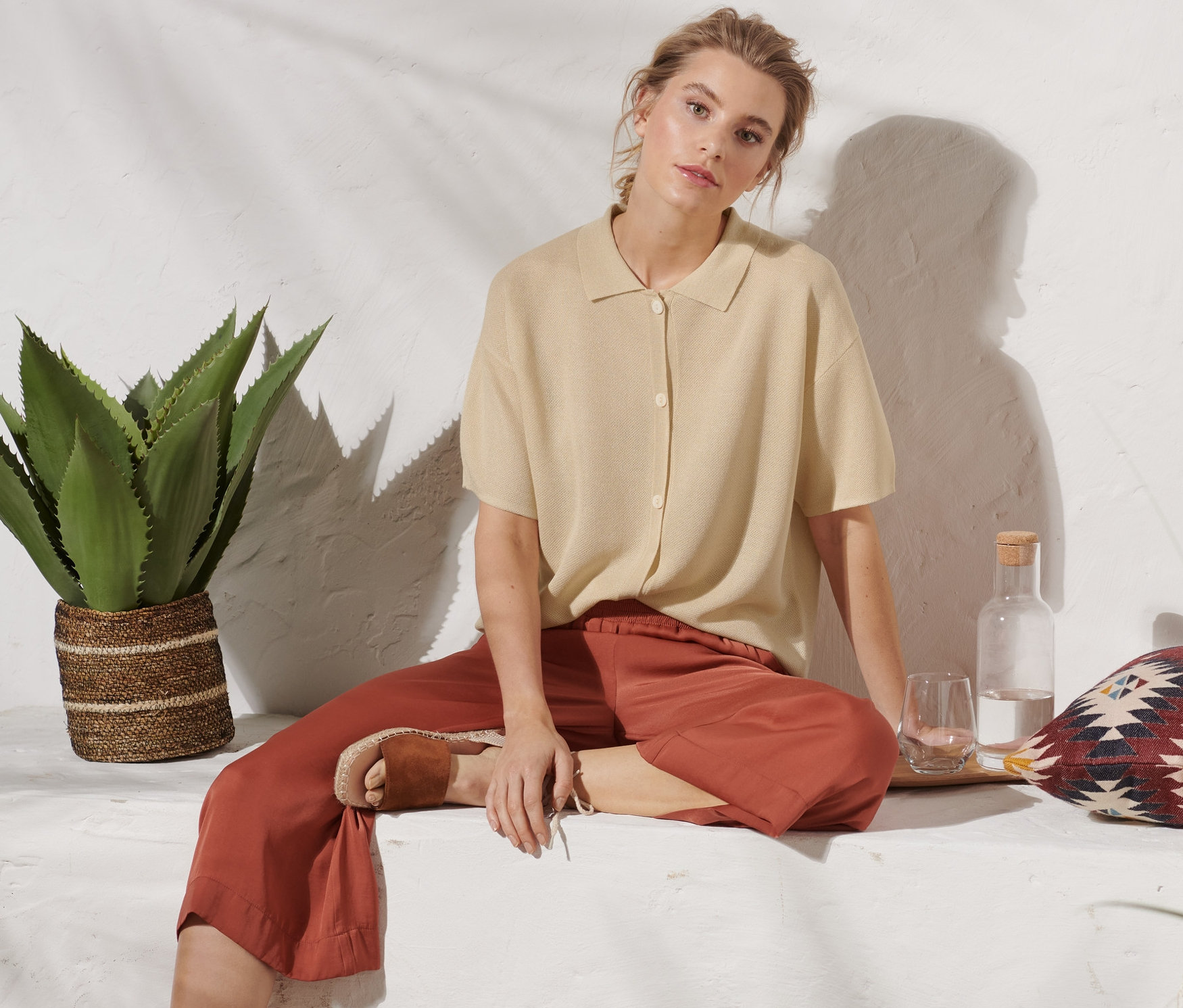 Eine Frau sitzt und trägt einen beigen Cardigan mit Kragen, braune Hosen und Sandalen.