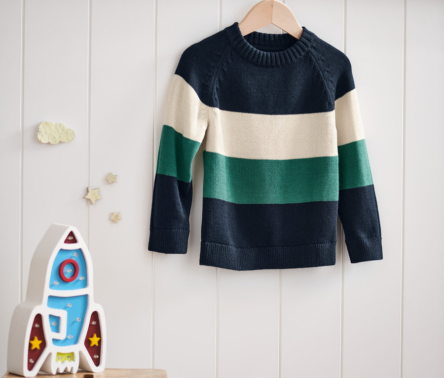 Ein gestreifter Strickpullover hängt auf einem Bügel an einer weißen Wand. Darunter steht eine Raketenlampe auf einem Hocker.