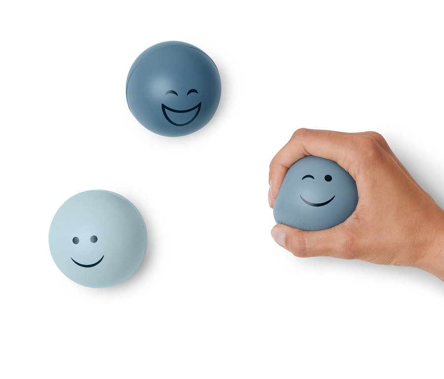 Eine Hand hält einen blauen Anti-Stressball mit Smiley, zwei weitere Anti-Stressbälle liegen daneben.