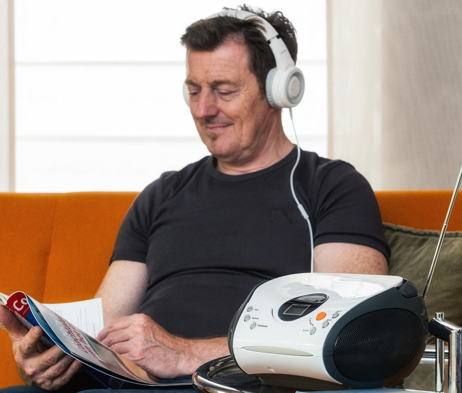 Ein Mann sitzt auf einem orangefarbenen Sofa, liest eine Broschüre und hört einen weißen Lenco »SCD-24 Boombox« CD-Player mit Radio und Kopfhöreranschluss.