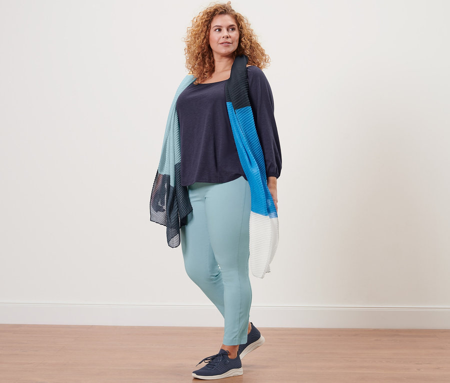 Frau steht und trägt ein dunkelblaues Blusenshirt mit 3/4-Arm, einen plissierten Schal und eine Stretchhose.