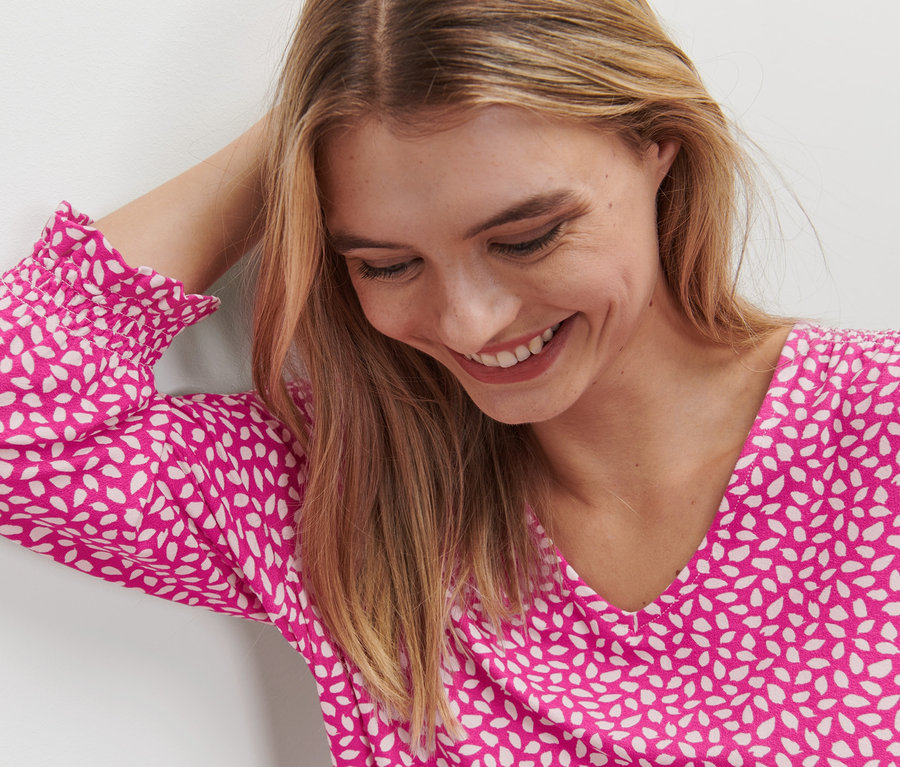 Frau lächelt und hat eine Hand im Haar. Sie trägt ein bedrucktes Blusenshirt in Pink.