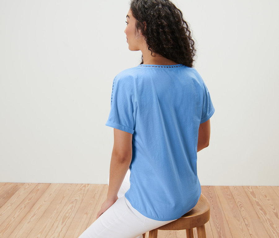 Eine Frau sitzt mit dem Rücken zur Kamera auf einem Hocker und trägt ein hellblaues Blusenshirt mit Crochet-Tape.