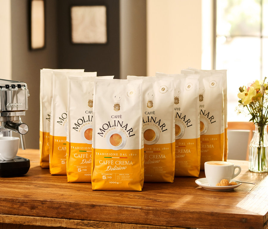 Auf einem Holztisch stehen eine Kaffeemaschine, acht Packungen Caffè Molinari Caffè Crema Delizioso und eine Blumenvase.