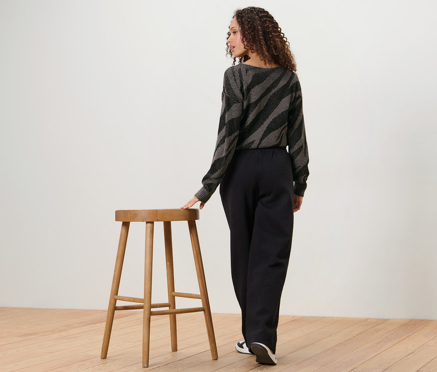 Frau steht mit dem Rücken zur Kamera und lehnt sich an einen Holzhocker. Sie trägt eine Sweathose und ein Oberteil mit Tiermuster.