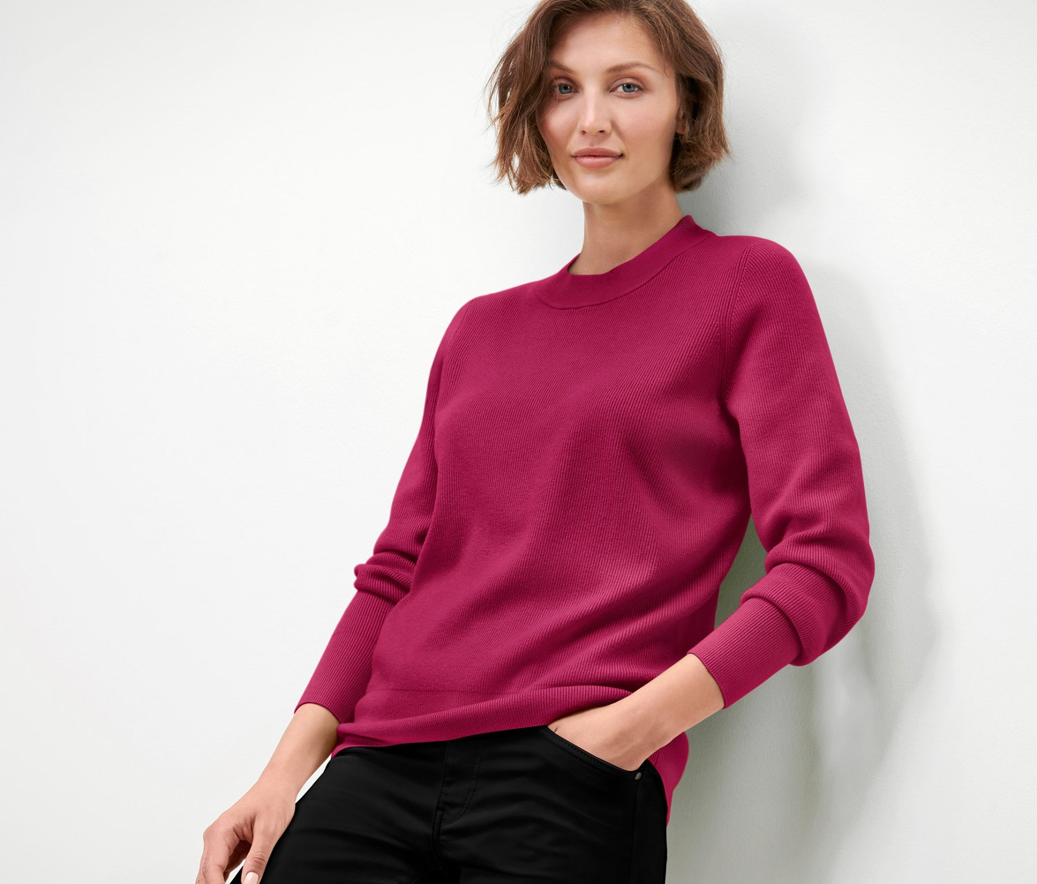 Eine Frau lehnt an einer Wand und trägt einen himbeerroten Feinstrickpullover und eine schwarze Hose.