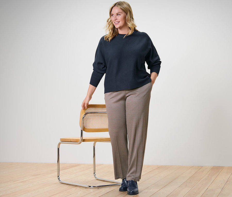 Frau steht neben einem Stuhl und trägt einen marineblauen Feinstrickpullover mit Fledermausarm und eine Jacquardhose.