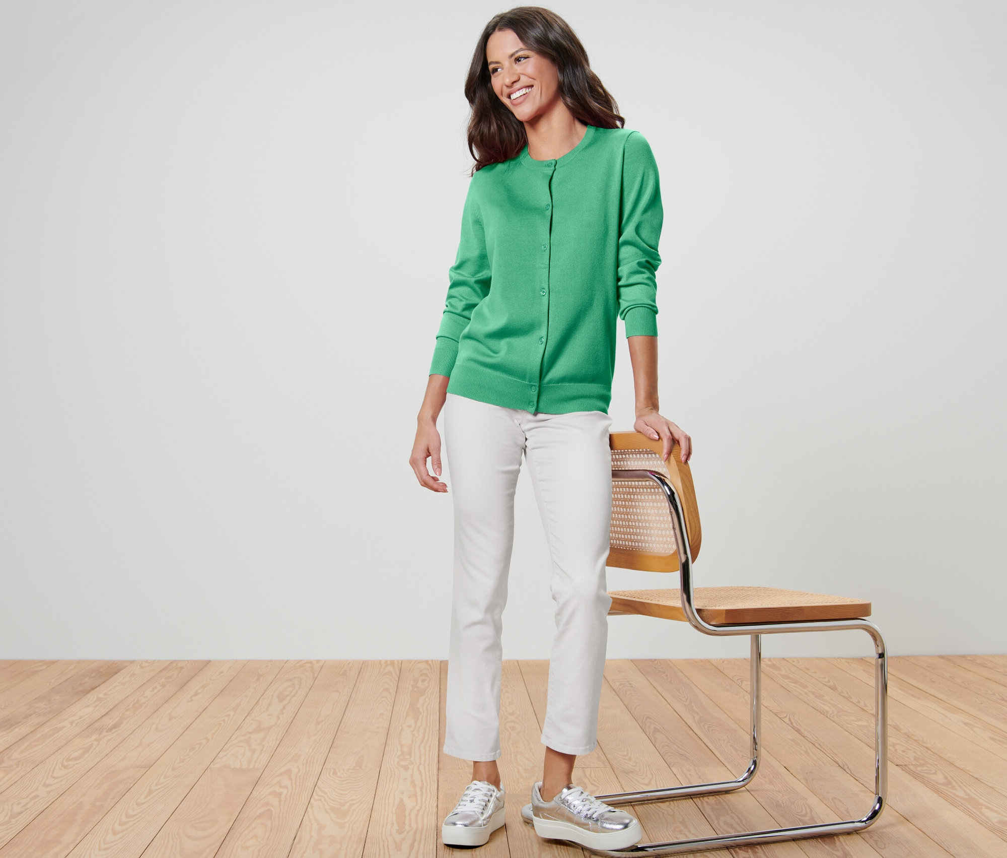 Frau steht an einem Stuhl gelehnt und trägt einen grünen Feinstrick-Cardigan, weiße Hosen und silberne Schuhe.