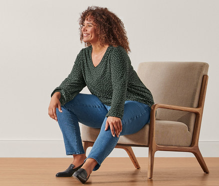 Eine Frau sitzt in einem Sessel und trägt ein gepunktetes Blusenshirt und eine Slimfit-Jeans.