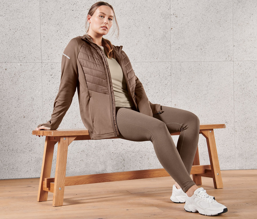 Eine Frau sitzt auf einer Bank und trägt eine braune Thermojacke mit Kapuze, ein taupefarbenes Langarm-Funktionsshirt und eine Premium-Seamless-Lauftight.
