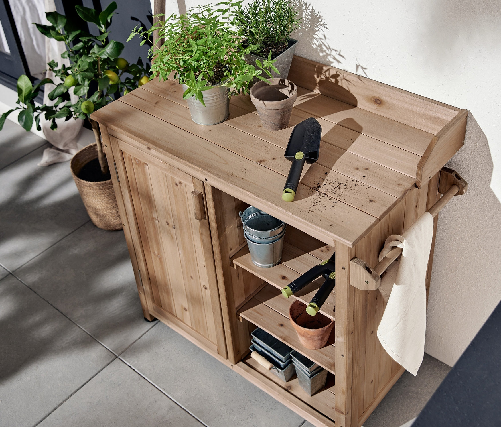 Ein Gartenschrank mit Handtuchhalter steht auf einer Terrasse, darauf liegen Blumen und Gartengeräte.