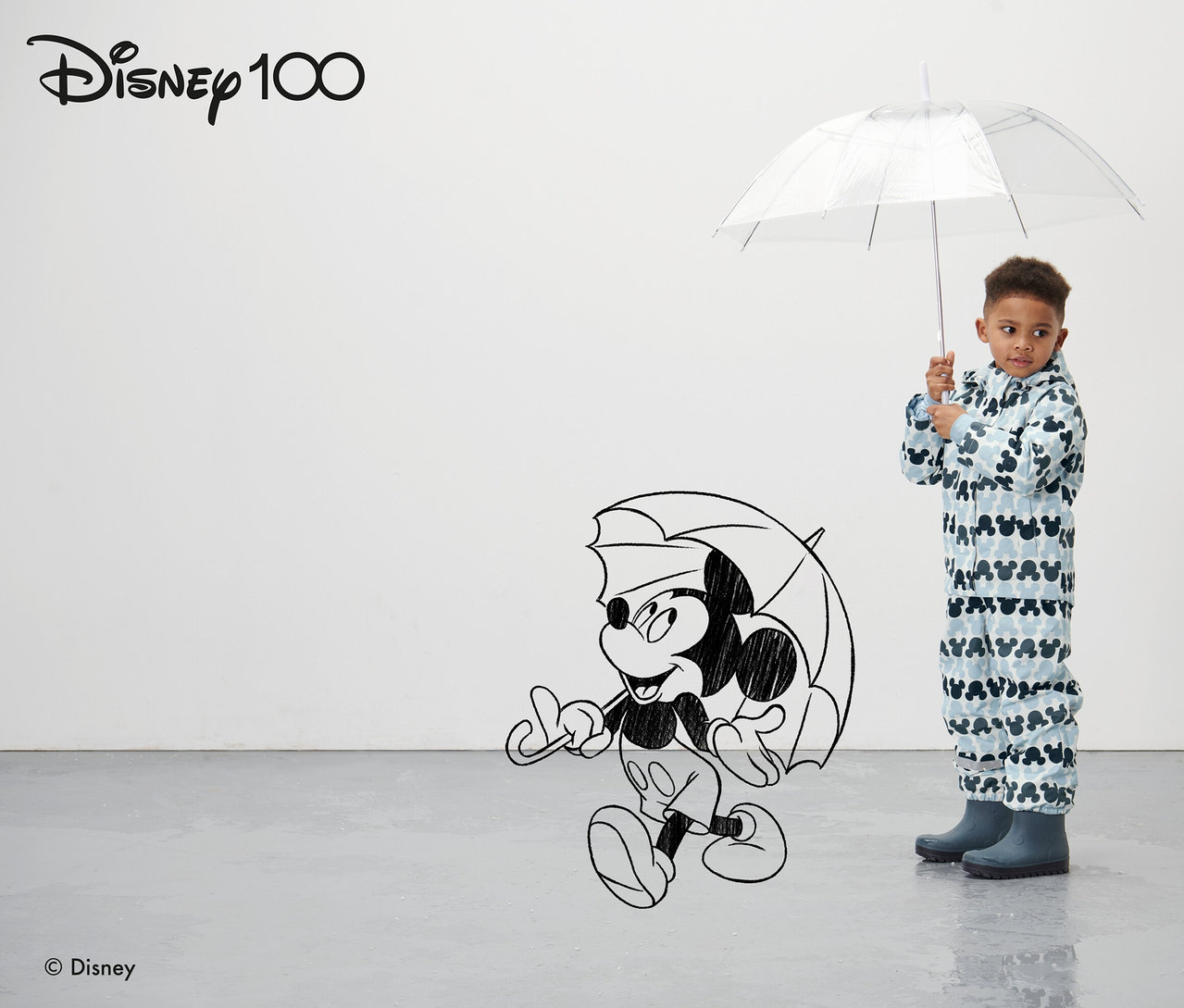 Ein Junge hält einen durchsichtigen Regenschirm. Er trägt eine blaue Kleinkind-Thermo-Regenjacke "Micky".