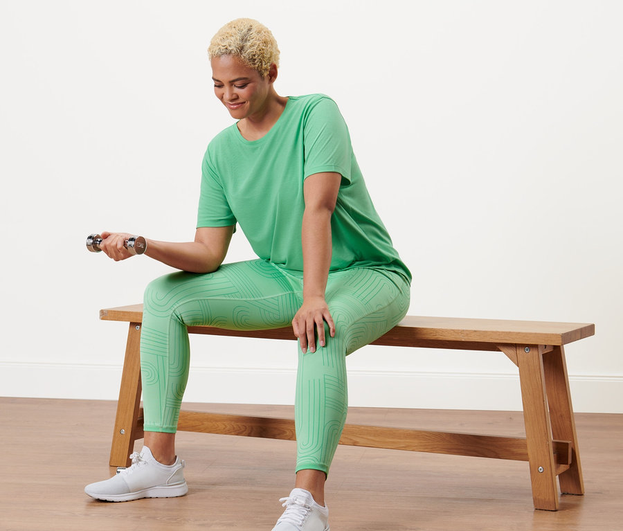 Frau sitzt auf einer Bank und trainiert mit einer Hantel in einem grünen Sportshirt und einer Wende-Sporttight.