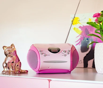 Auf einer rosa Kommode stehen eine rosa Lenco SCD-24 Boombox, eine goldene Katzenstatue, eine Blume in einem weißen Topf und eine Kerze.