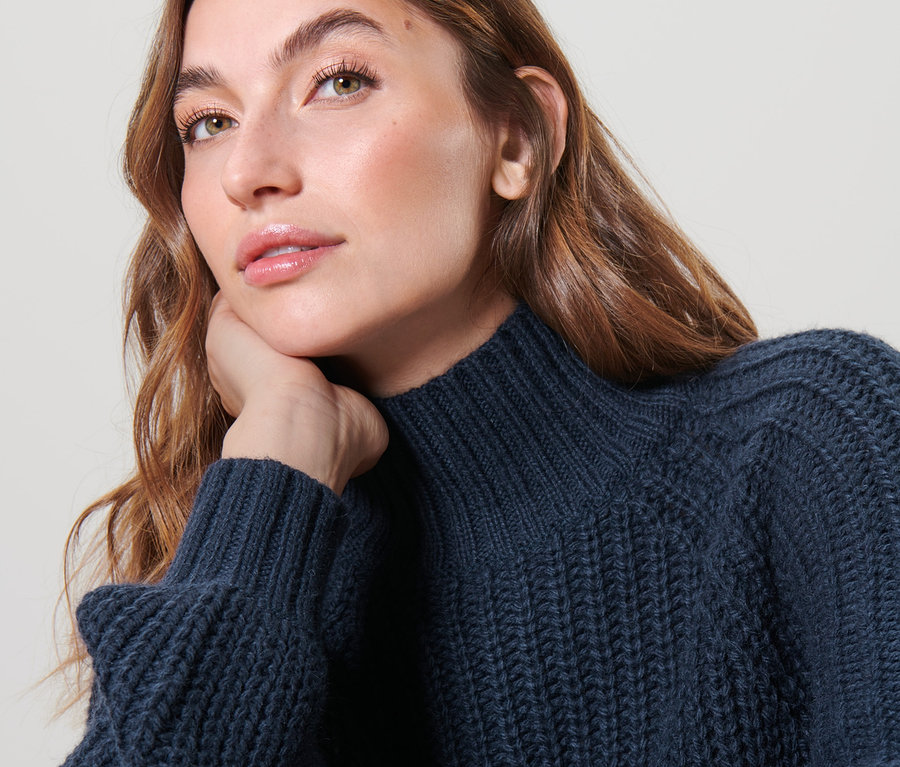 Frau mit braunen Haaren, die ihr Kinn auf ihre Hand stützt und einen dunkelblauen Strickpullover mit Stehkragen trägt.