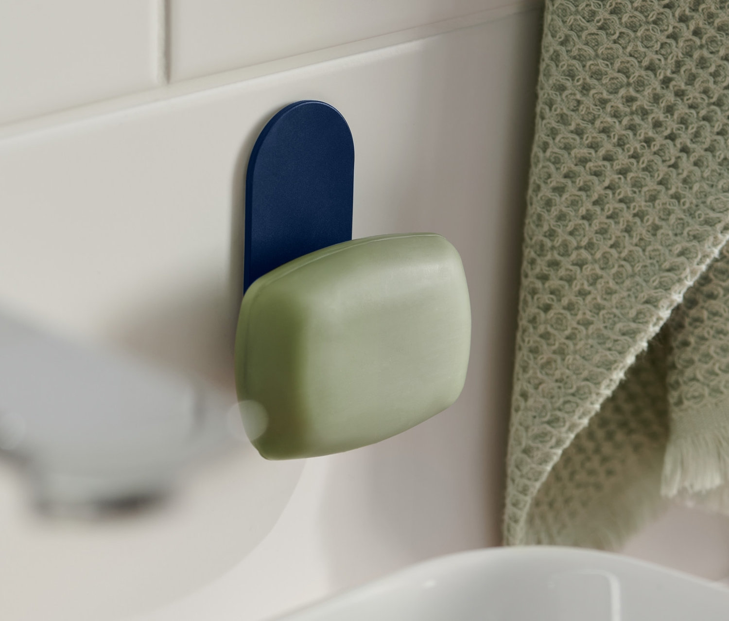Eine grüne Seife hängt an einem blauen Seifenhalter mit Magnet, der an weißen Fliesen neben einem Waschbecken befestigt ist. Rechts hängt ein grünes Handtuch.