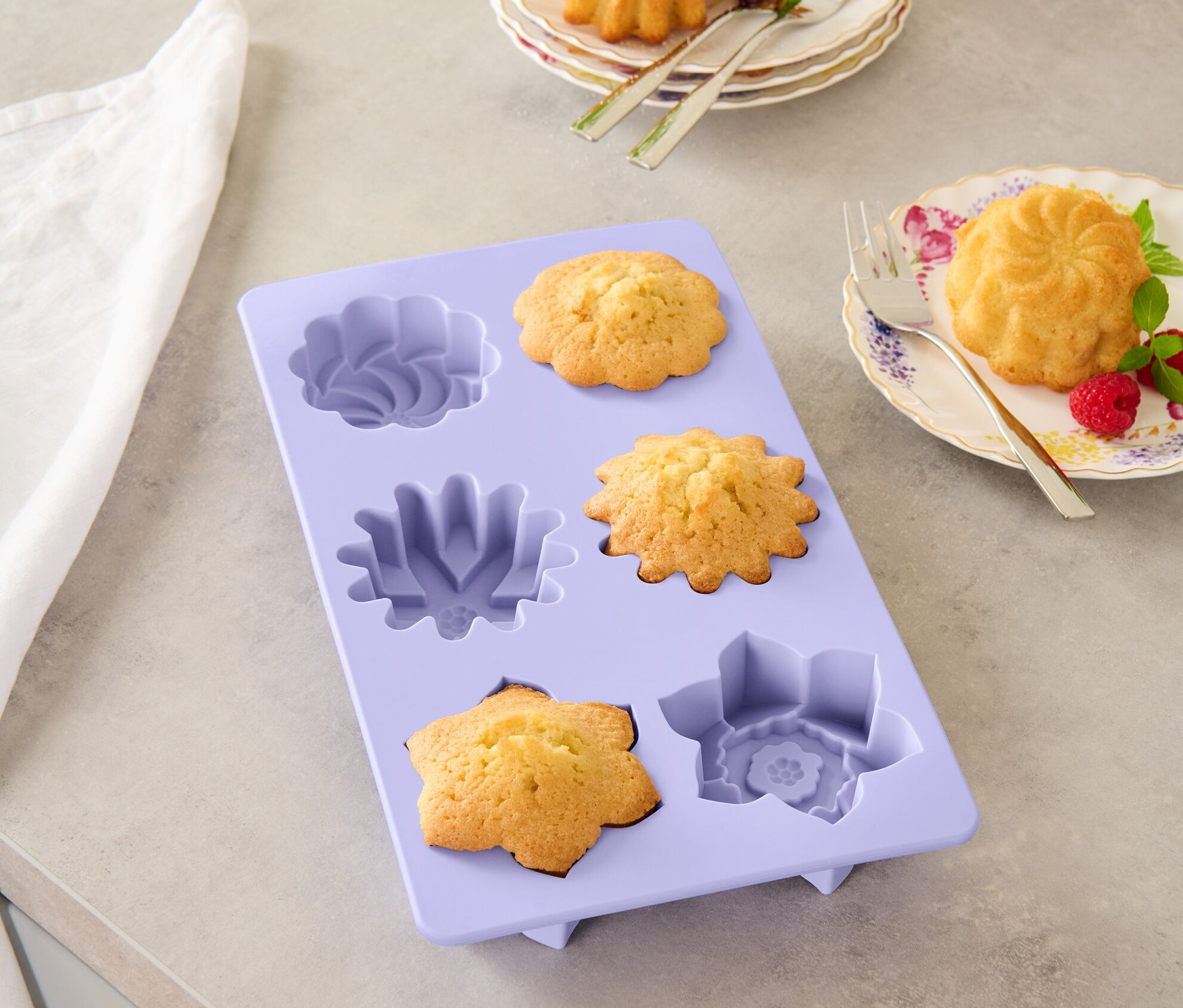 Lila Blüten-Backform mit gebackenen Küchlein, zwei Küchlein auf Tellern mit Gabeln.