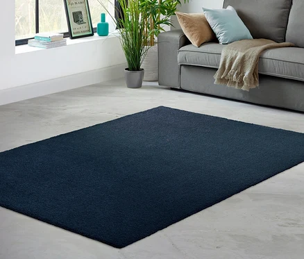 Im Vordergrund befindet sich ein blauer Teppich »Mellow«, im Hintergrund ein Wohnzimmer mit Sofa, Fenster und Pflanzen.