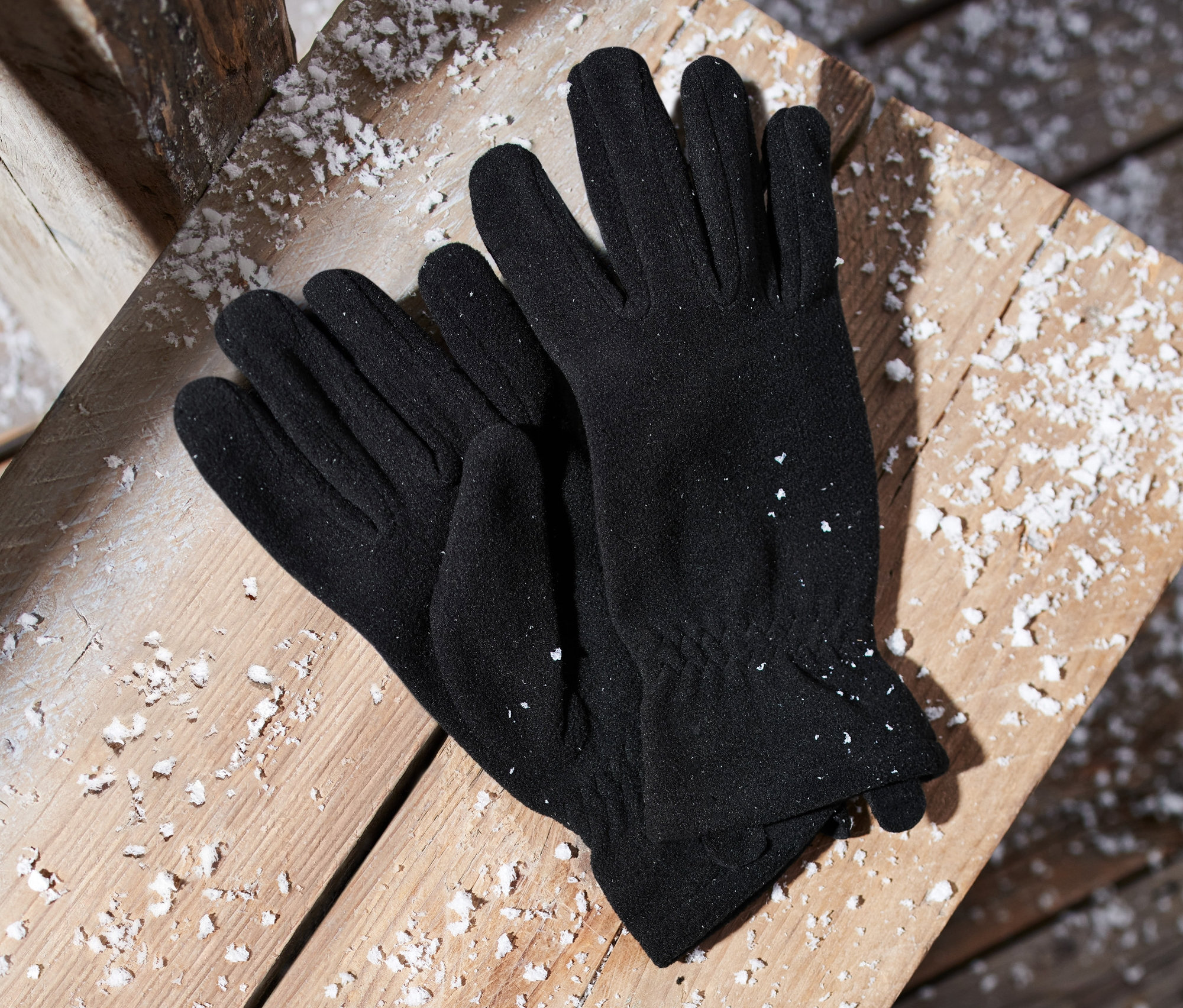 Schwarze Microfleece-Handschuhe liegen auf einer mit Schnee bedeckten Holzfläche.