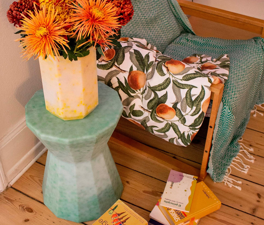 Blumenstrauß orangefarbener Chrysanthemen in gelber Vase auf grünem Beistelltisch neben Sessel mit Überwurf und Kissen mit Pfirsichmuster.