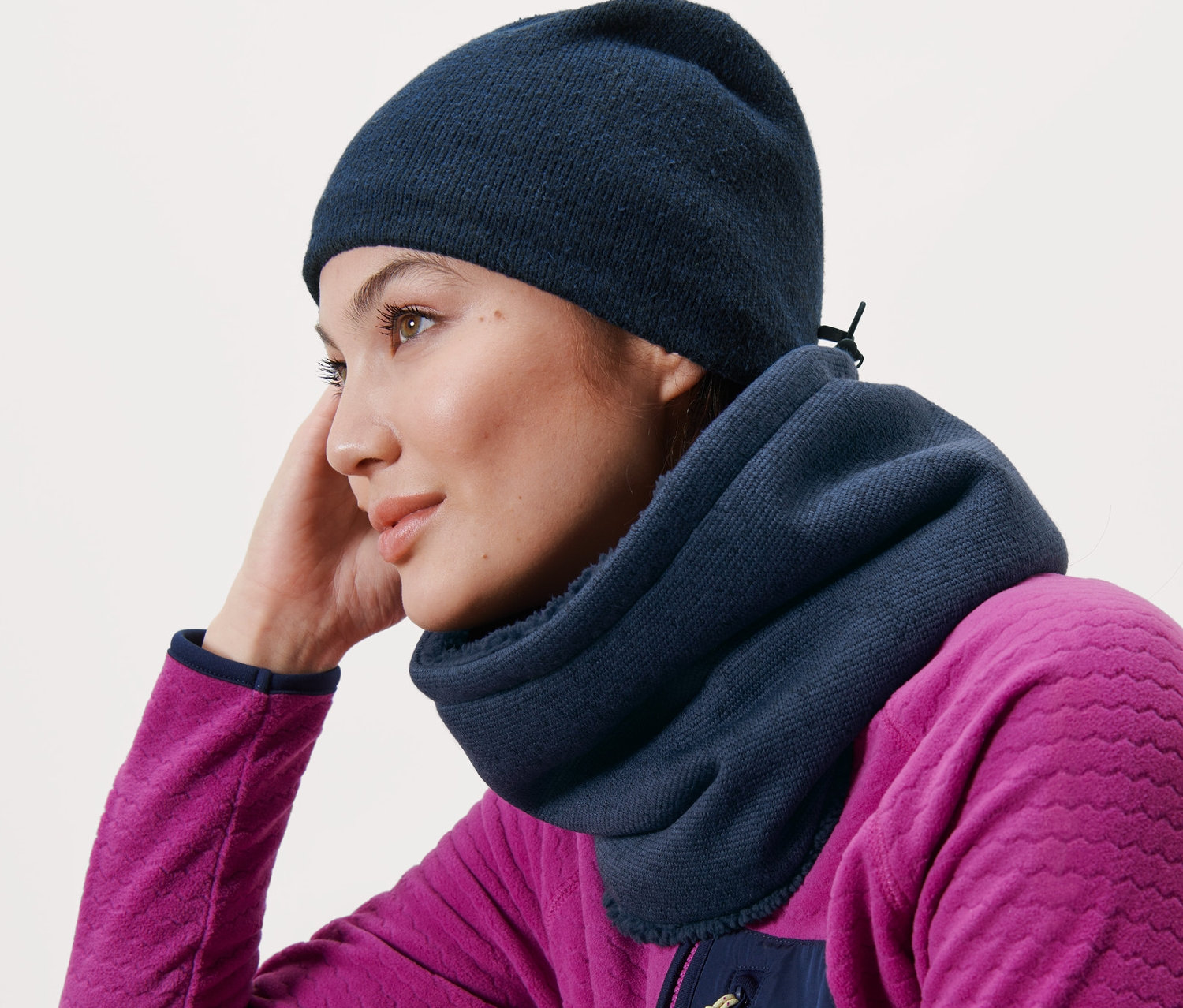 Eine Frau, die sich an eine Holzbank lehnt, trägt einen blauen Strickfleece-Schlauchschal und eine blaue Mütze.