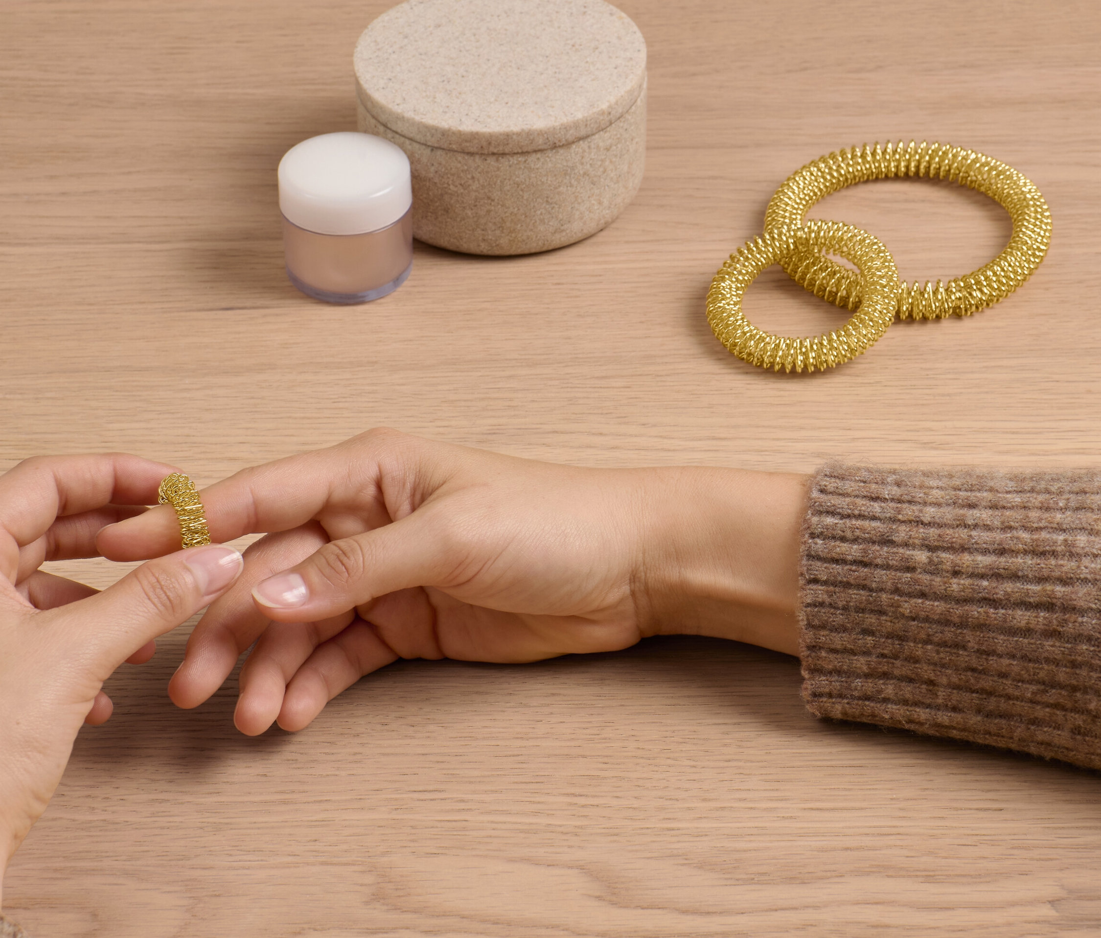 Frau schiebt goldenen Akupressurring vom Set auf ihren Finger. Zwei weitere Ringe und eine Dose liegen auf dem Tisch.