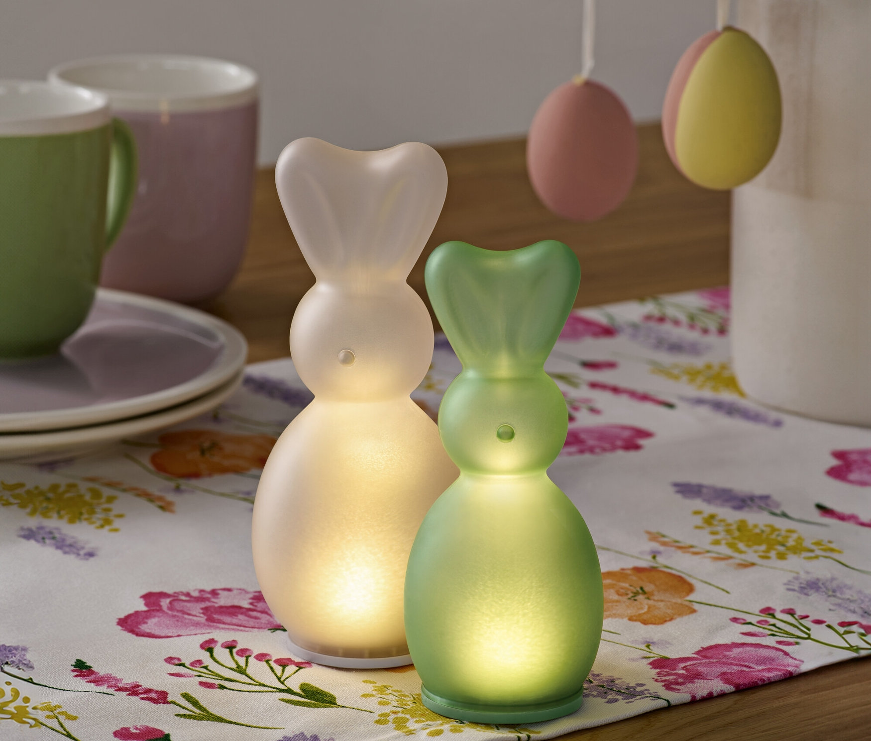 Zwei LED-Hasen stehen auf einem floralen Tischläufer, im Hintergrund Tassen und Ostereier.
