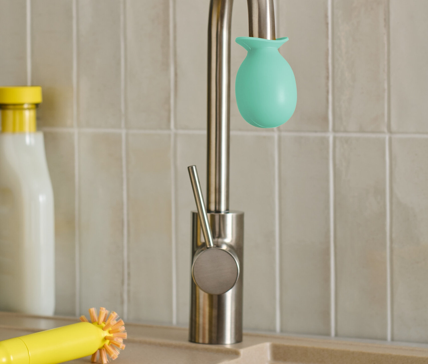 Auf einem Wasserhahn sitzt ein grüner Wasserhahn Entkalker-Aufsatz. Daneben liegen eine gelbe Bürste und ein Putzlappen.