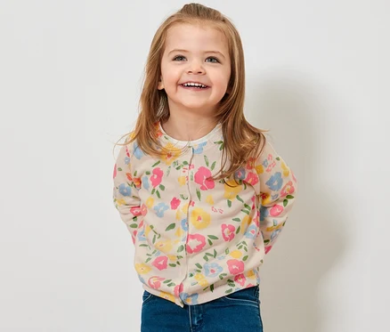 Lächelndes Mädchen trägt eine Kinder-Strickjacke mit Blumenmuster und Jeans.