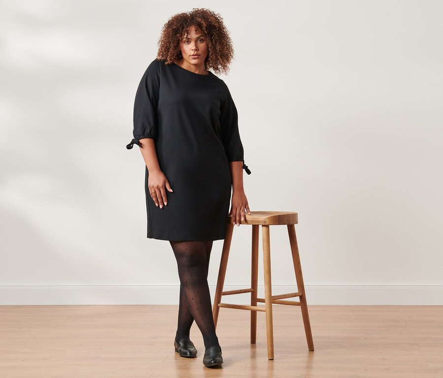 Eine Frau posiert neben einem Holzhocker, sie trägt ein schwarzes Jerseykleid mit Schleifendetail und eine gepunktete Feinstrumpfhose.