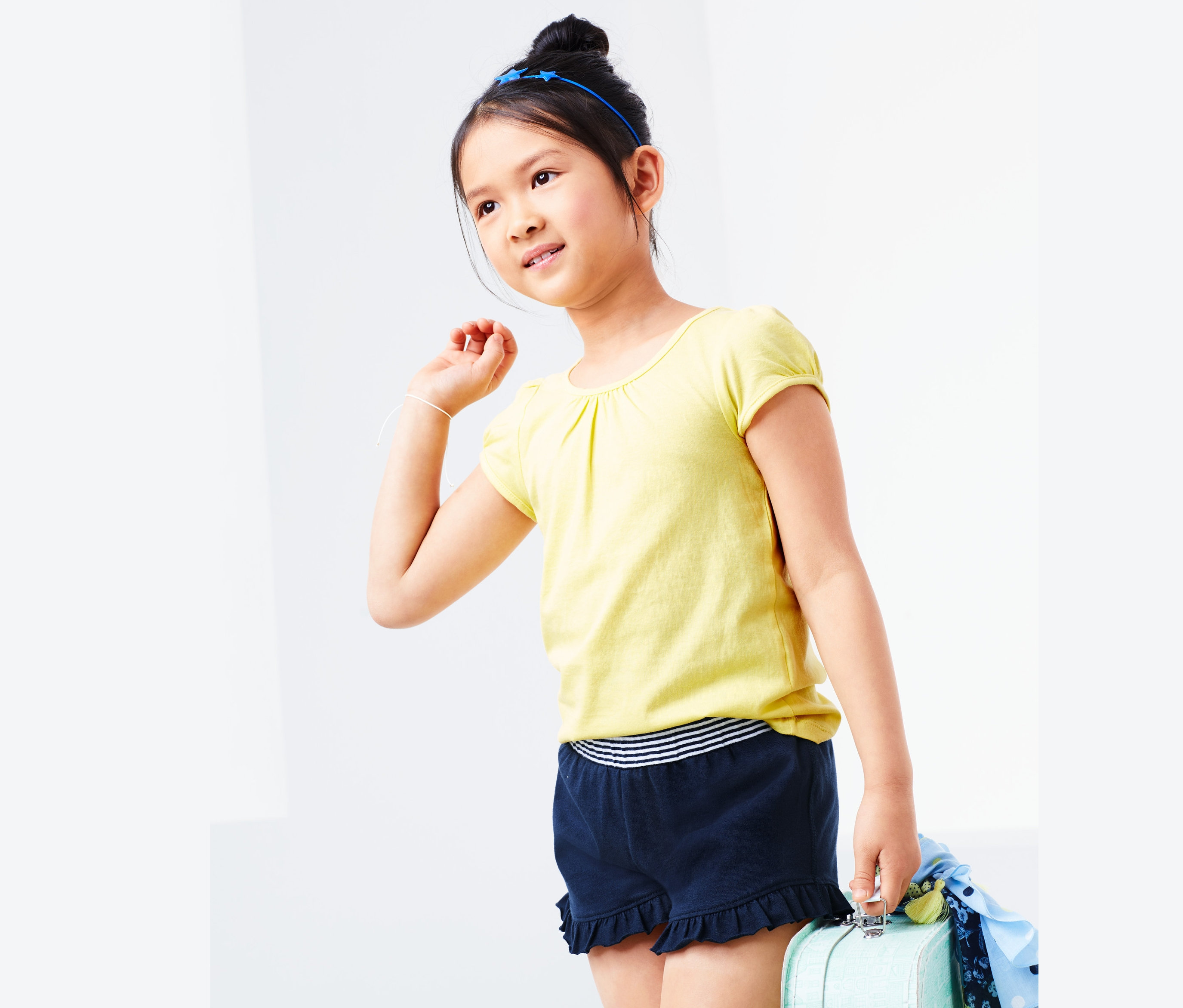 Mädchen in gelbem T-Shirt und dunkelblauen Jersey-Shorts hält einen blauen Koffer.