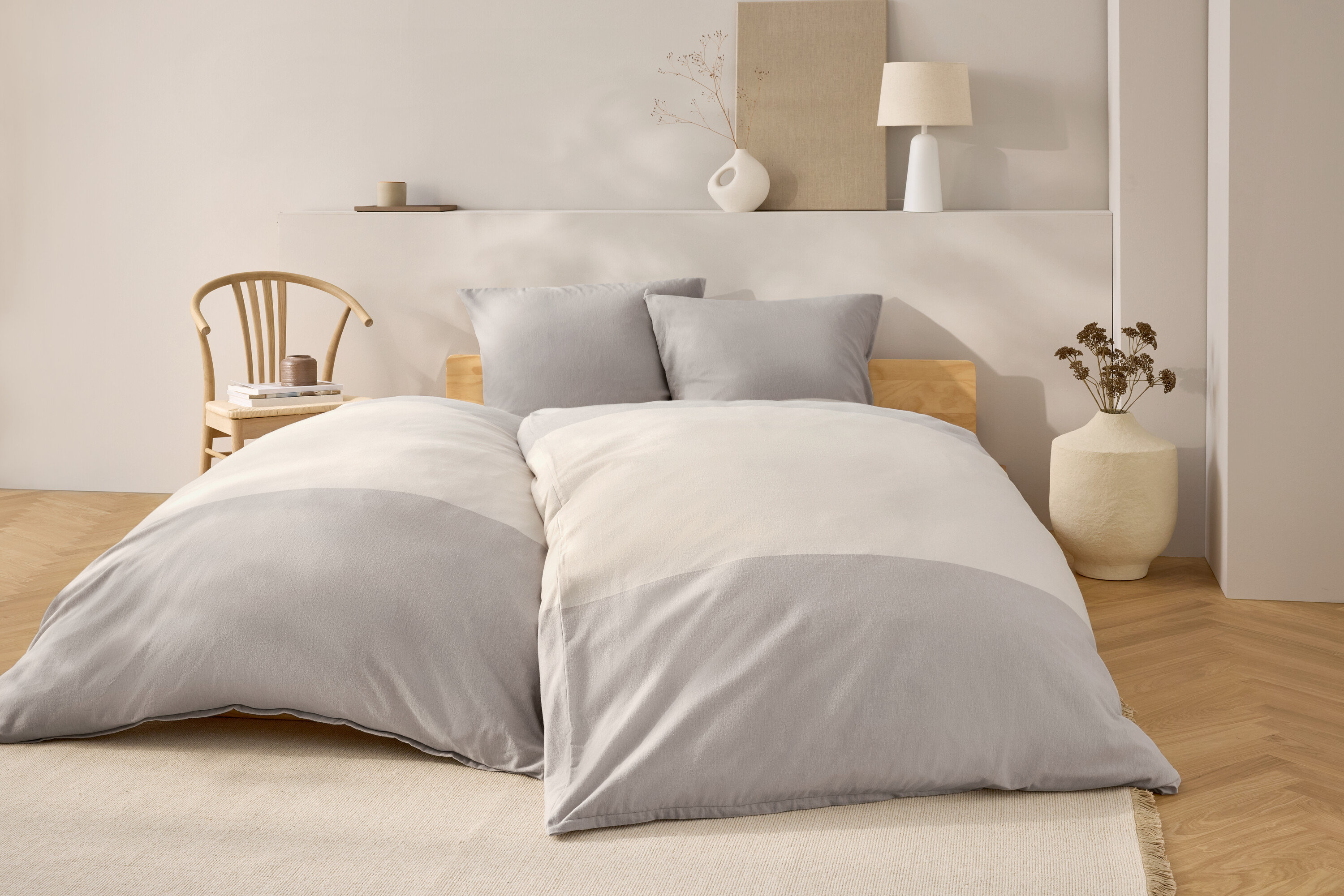 Schlafzimmer mit Bett mit grau-weisser Bettwäsche, zwei grauen Kissen und einem hölzernen Kopfteil. Im Hintergrund ein Holzstuhl und Dekoration.