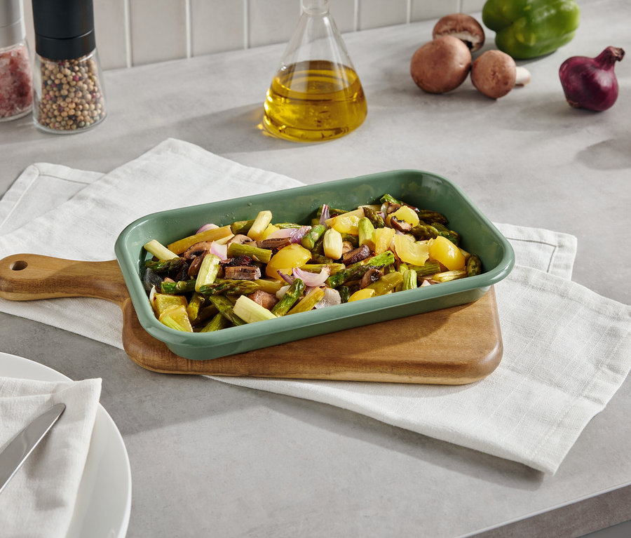 Grüne Auflaufform mit gebackenem Gemüse auf einem Brett mit Serviette. Im Hintergrund befinden sich Pilze, Paprika, Zwiebeln und Gewürze.