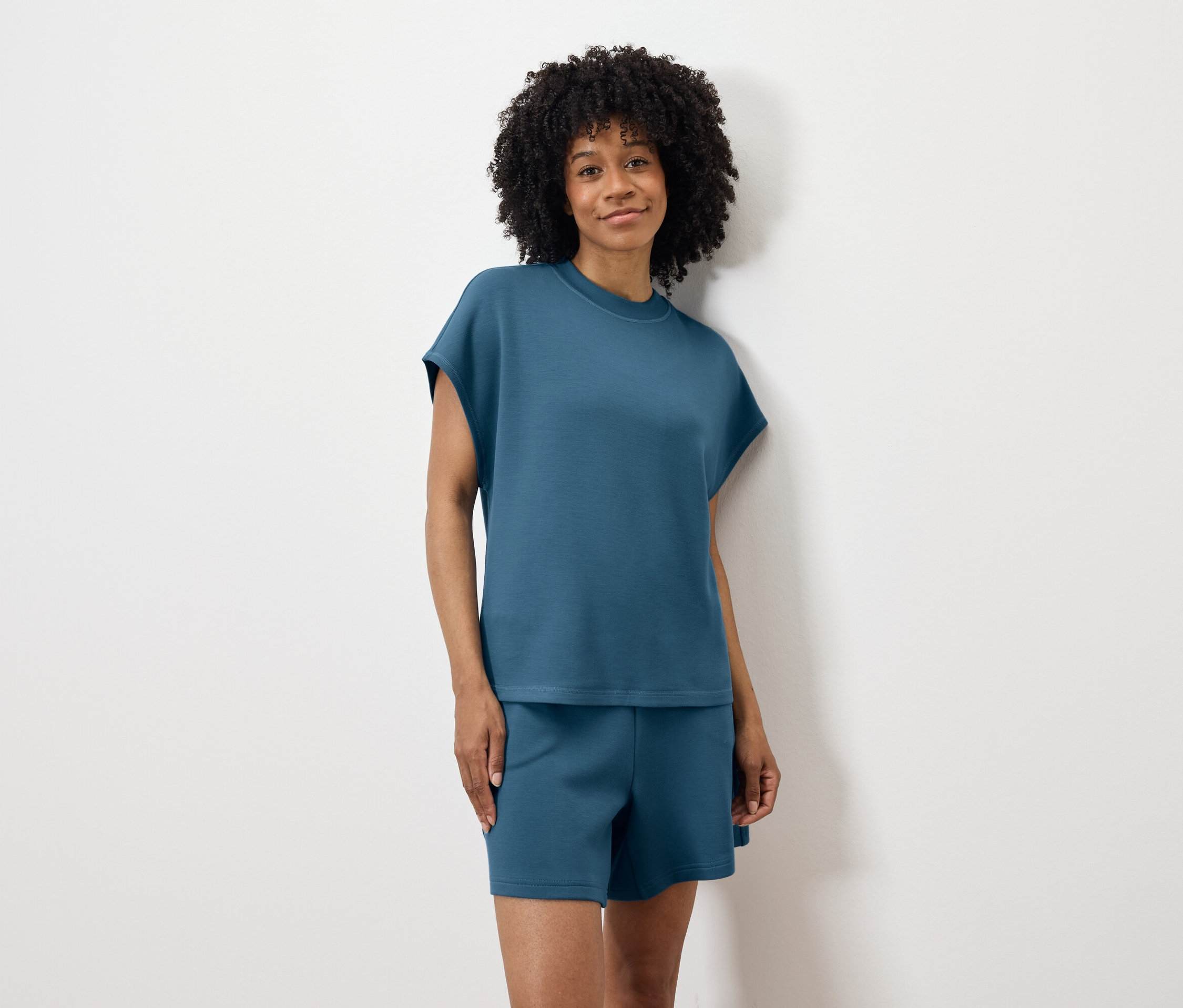 Frau mit lockigem Haar, in blauem T-Shirt und Shorts, steht vor einer weißen Wand.