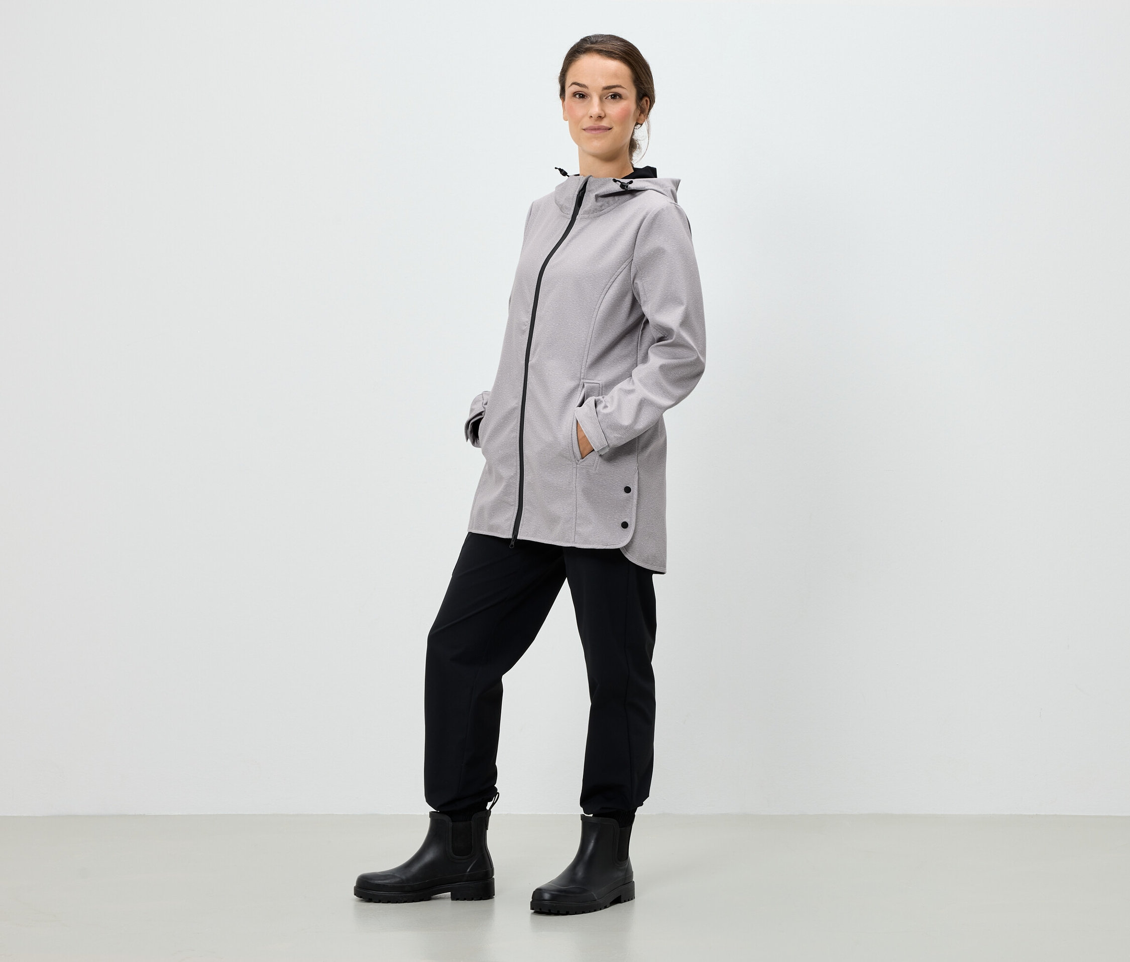 Eine Frau steht mit den Händen in den Taschen einer grauen Kapuzenjacke, schwarzen Hosen und schwarzen Stiefeln.