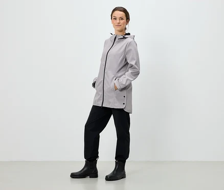 Eine Frau steht mit den Händen in den Taschen einer grauen Kapuzenjacke, schwarzen Hosen und schwarzen Stiefeln.