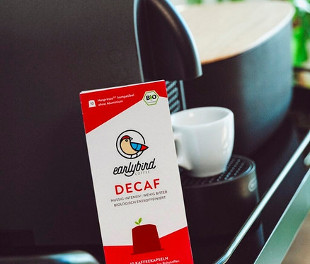 Eine Packung Earlybird Decaf Kaffeekapseln steht neben einer Kaffeemaschine und einer Tasse.