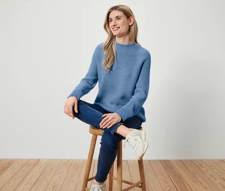 Frau sitzt auf einem Holzhocker, trägt einen blauen Pullover, dunkelblaue Jeans und weiße Turnschuhe.